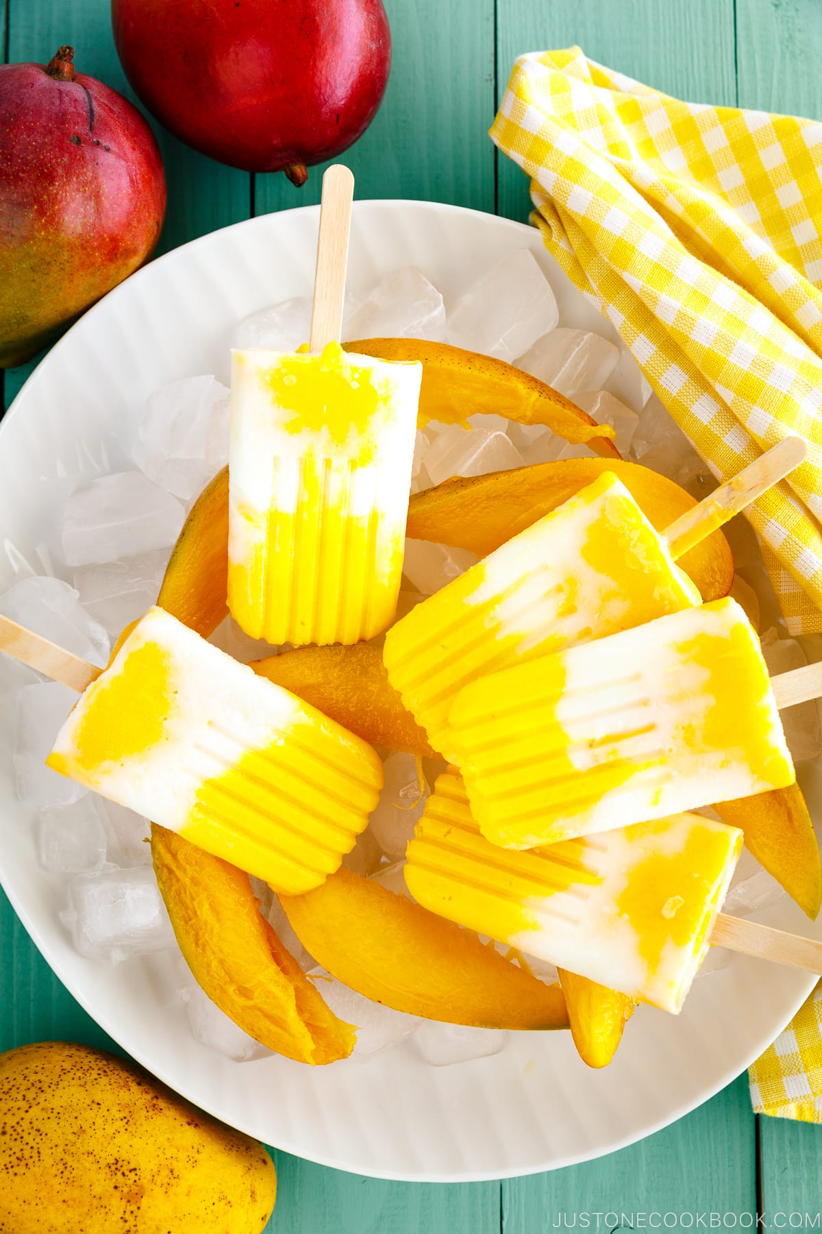 A white dish containing Mango Lassi Popsicles over ice cubes and fresh sliced mangos.