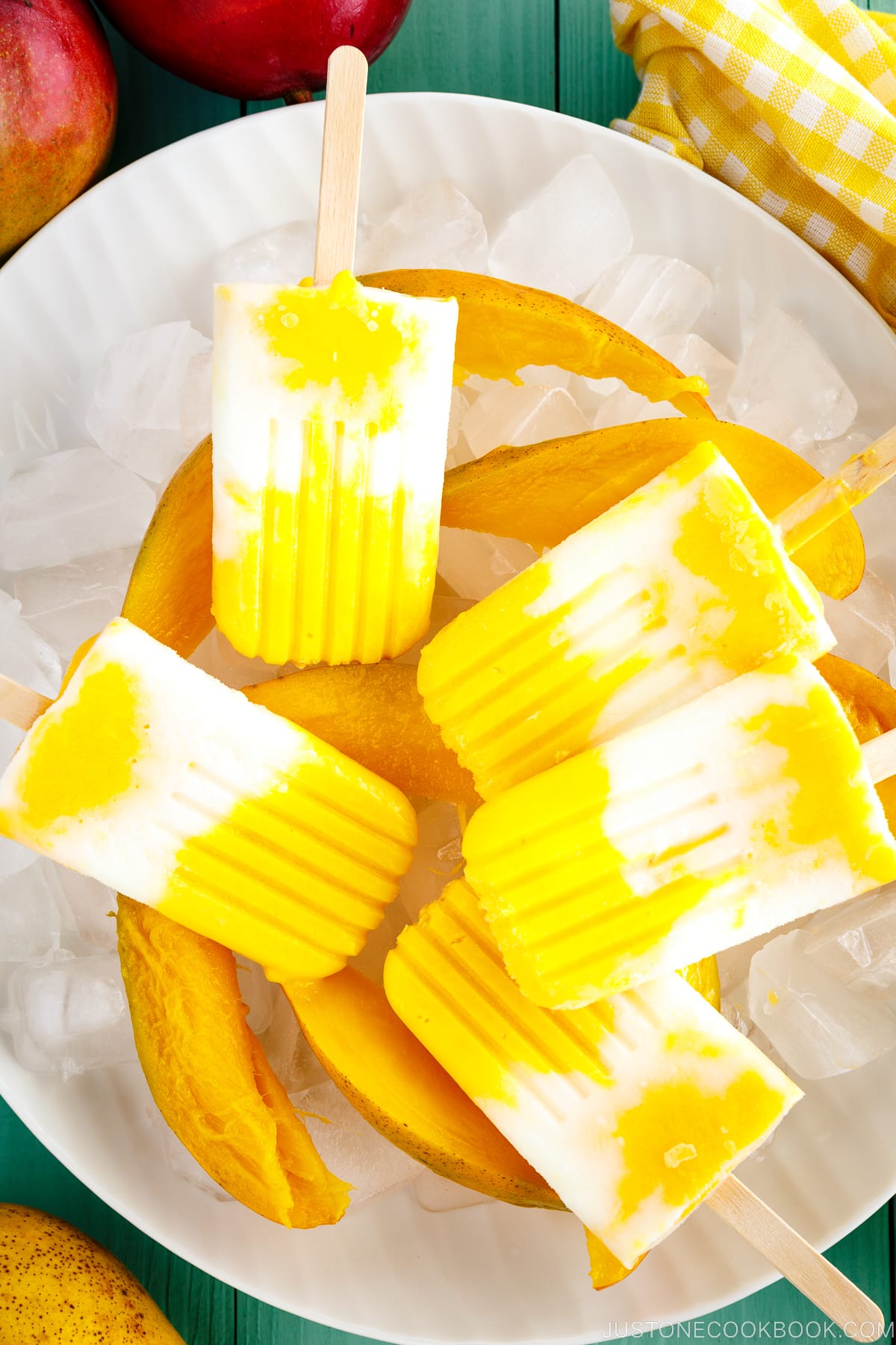 A white dish containing Mango Lassi Popsicles over ice cubes and fresh sliced mangos.