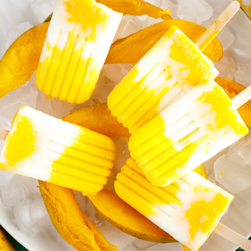 A white dish containing Mango Lassi Popsicles over ice cubes and fresh sliced mangos.