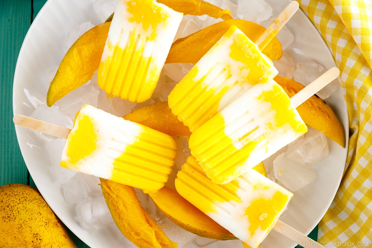 A white dish containing Mango Lassi Popsicles over ice cubes and fresh sliced mangos.