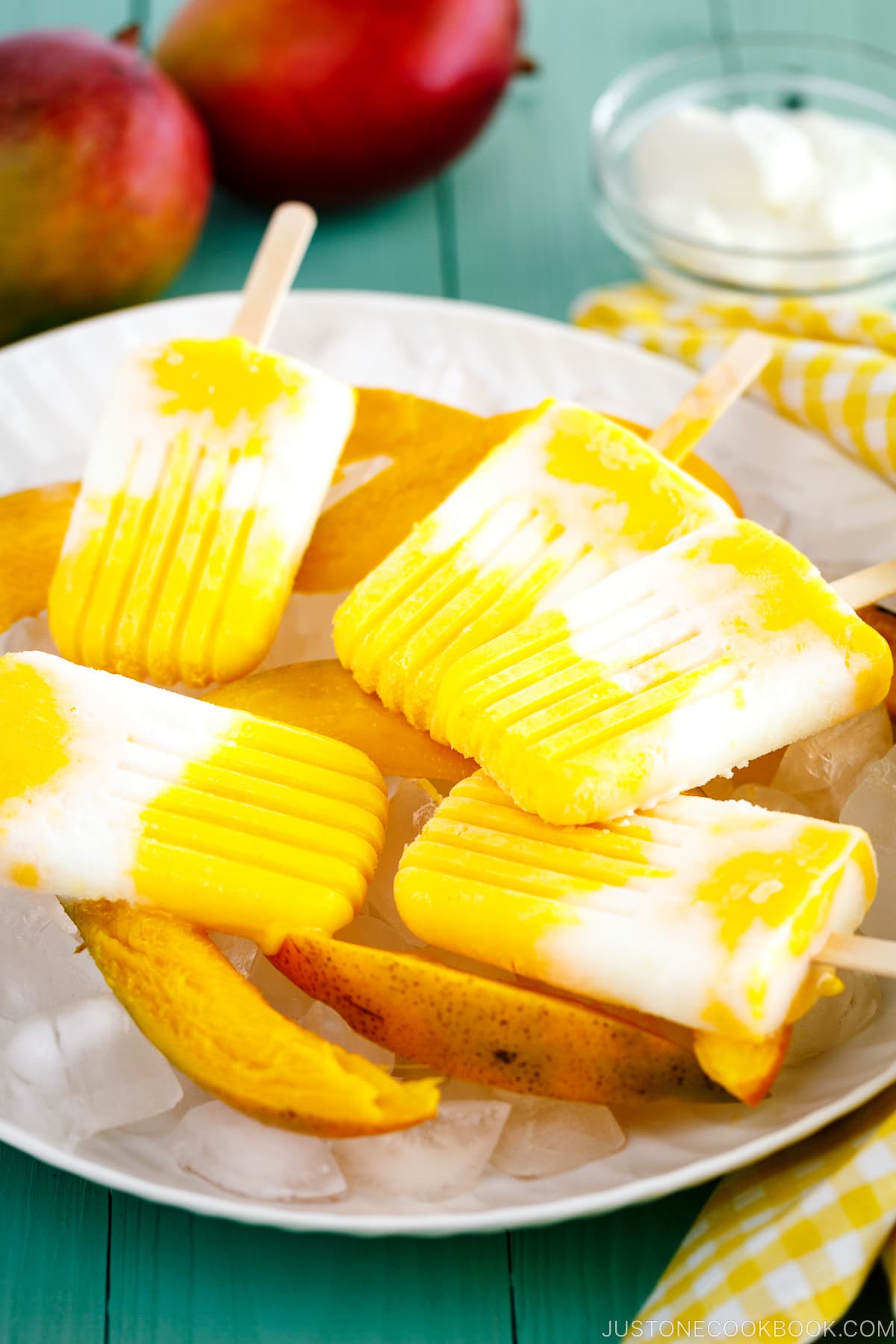 A white dish containing Mango Lassi Popsicles over ice cubes and fresh sliced mangos.