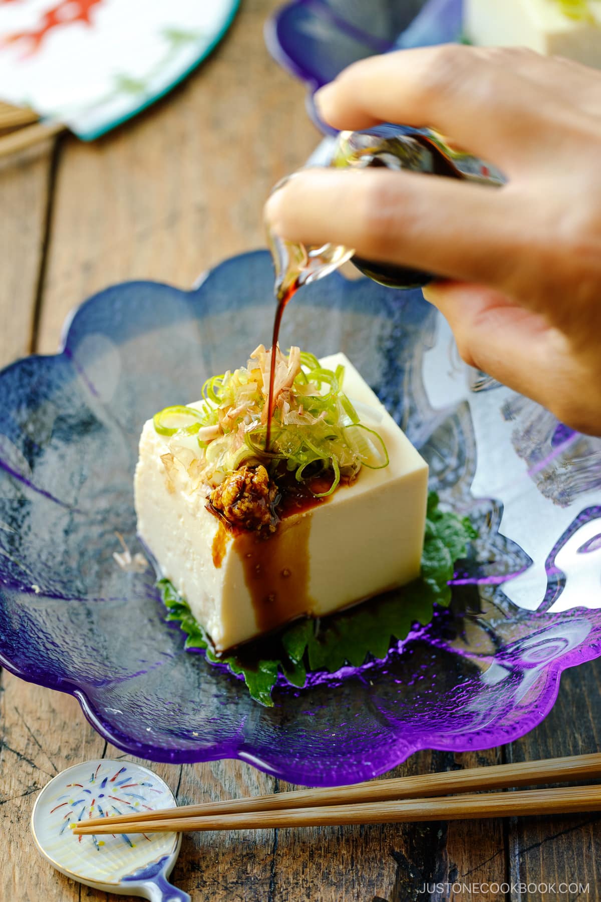 A glassware containing Japanese cold tofu topped with green onion, grated ginger, and katsuobushi and drizzled with soy sauce.