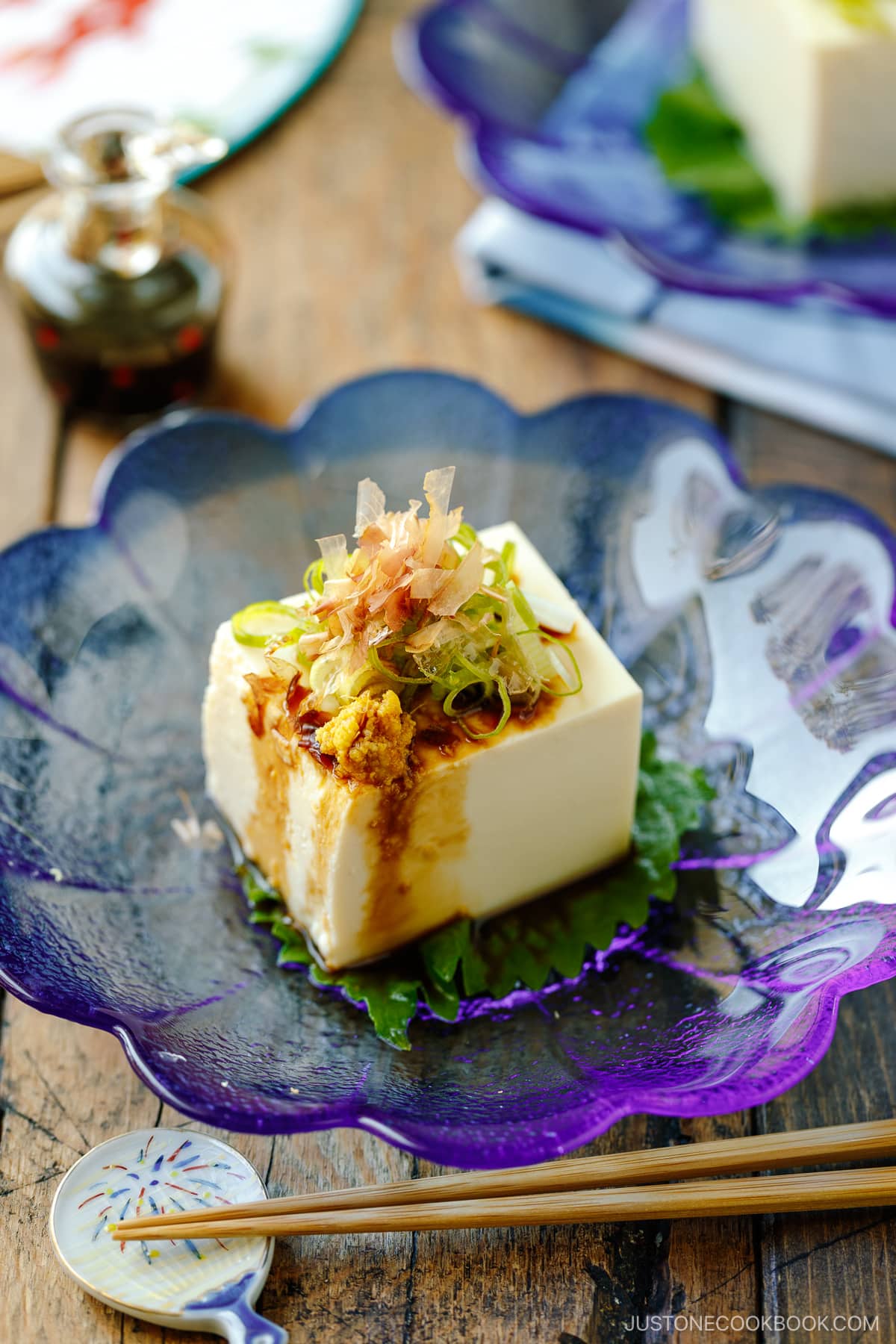 A glassware containing Japanese cold tofu topped with green onion, grated ginger, and katsuobushi and drizzled with soy sauce.