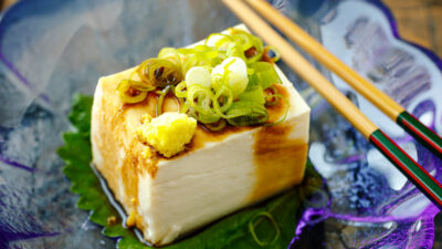 A glassware containing Japanese cold tofu topped with green onion and grated ginger and drizzled with soy sauce.