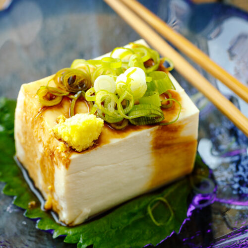 A glassware containing Japanese cold tofu topped with green onion and grated ginger and drizzled with soy sauce.