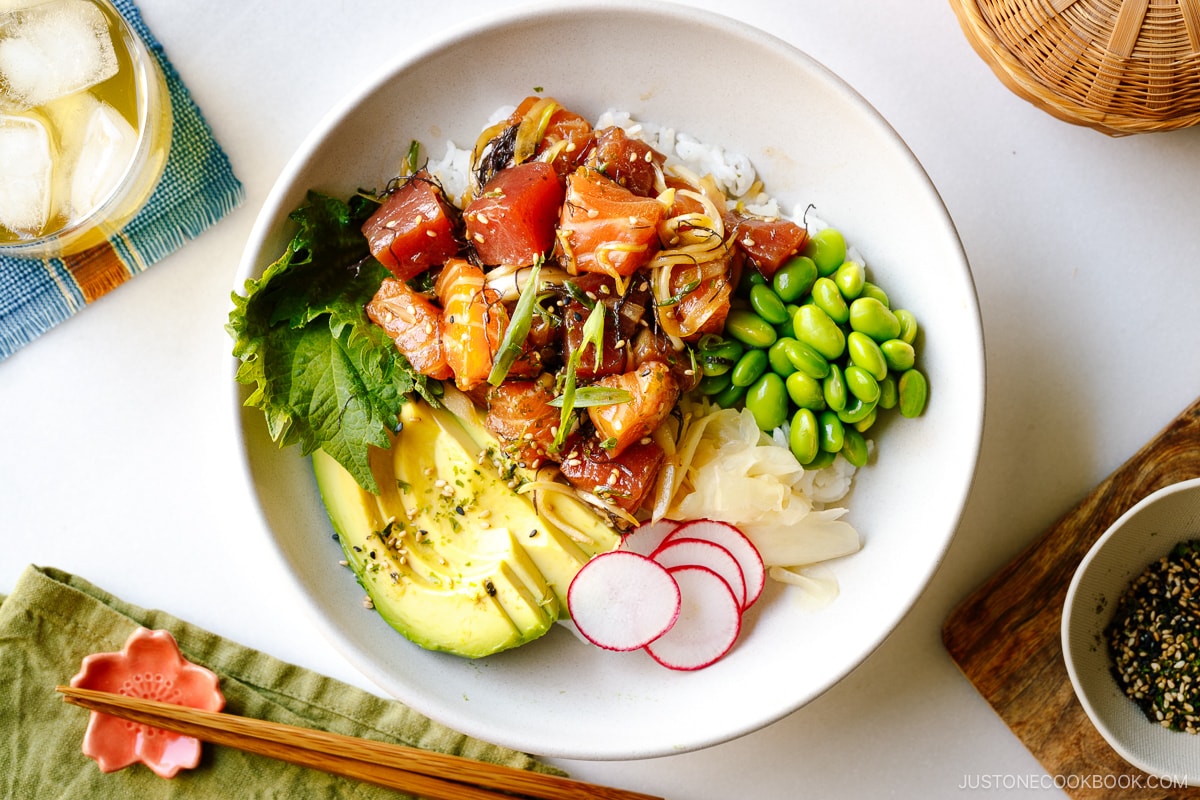 A bowl containing ahi tuna and salmon poke along with edamame, avocado, sushi ginger, and radish over bed of steamed rice.