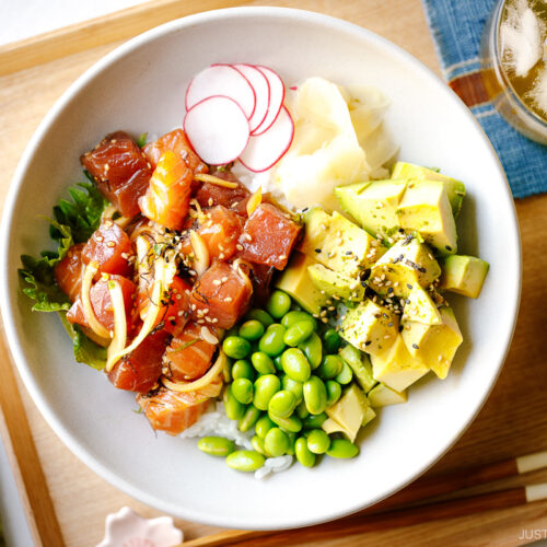 A bowl containing ahi tuna and salmon poke along with edamame, avocado, sushi ginger, and radish over bed of steamed rice.