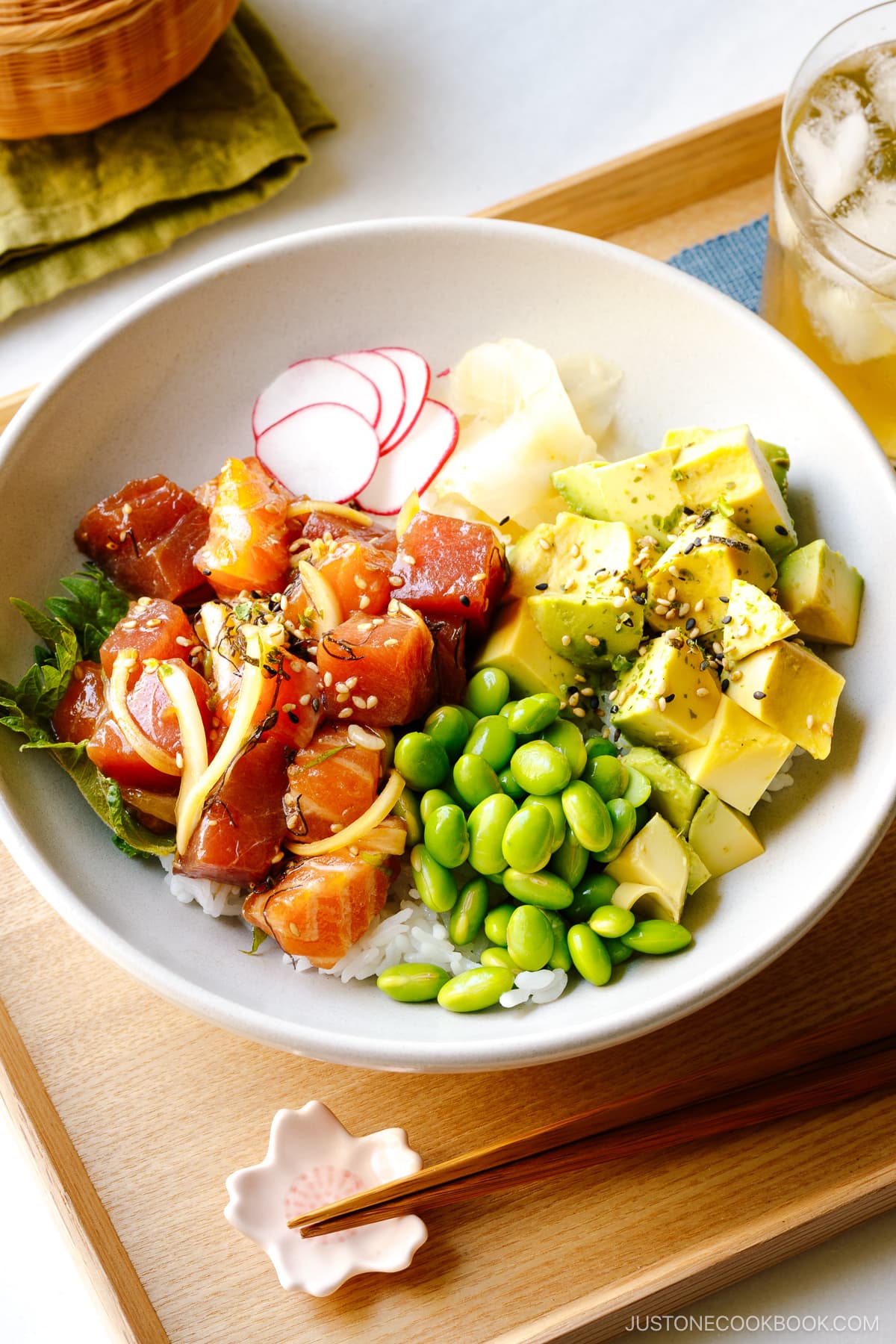 A bowl containing ahi tuna and salmon poke along with edamame, avocado, sushi ginger, and radish over bed of steamed rice.