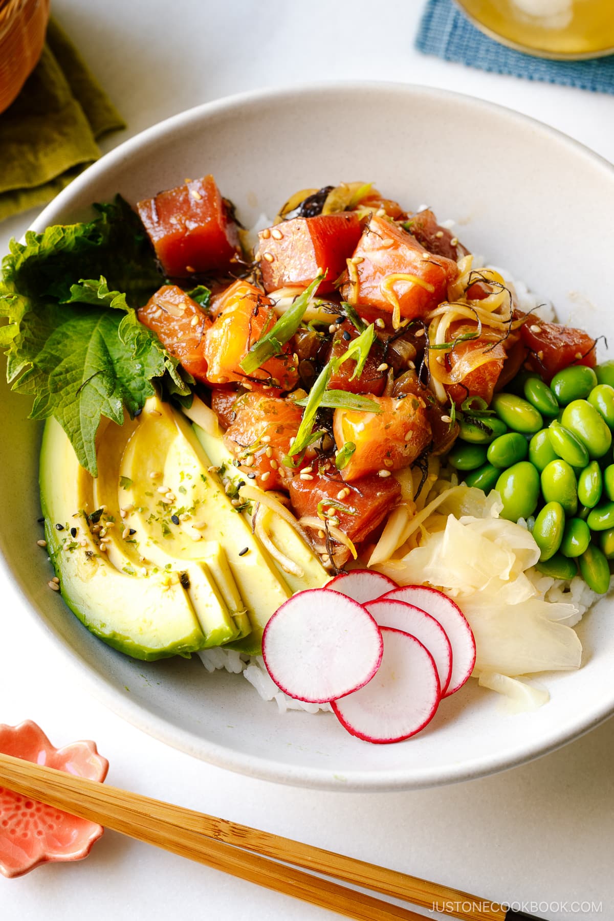 A bowl containing ahi tuna and salmon poke along with edamame, avocado, sushi ginger, and radish over bed of steamed rice.