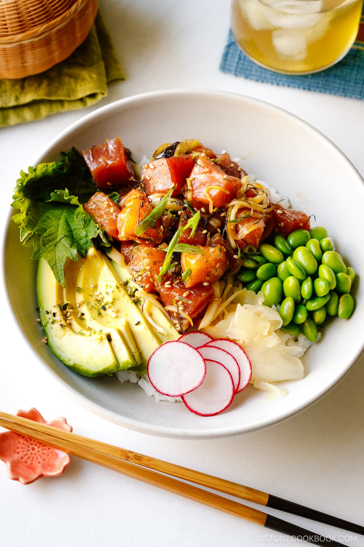 A bowl containing ahi tuna and salmon poke along with edamame, avocado, sushi ginger, and radish over bed of steamed rice.