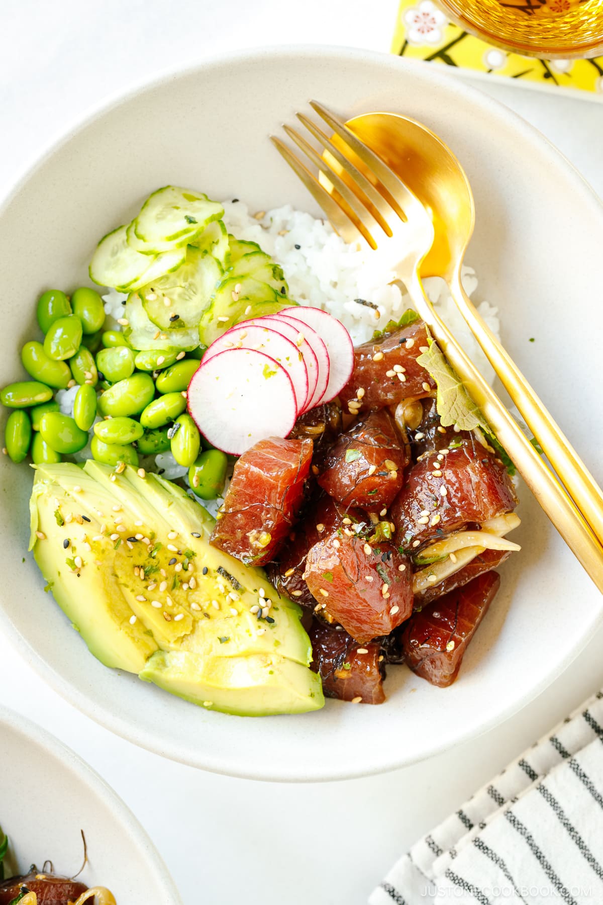 Ahi Tuna Poke Bowl containing soy marinated raw tuna, avocado slices, edamame, and cucumber slices over a bed of steamed rice.