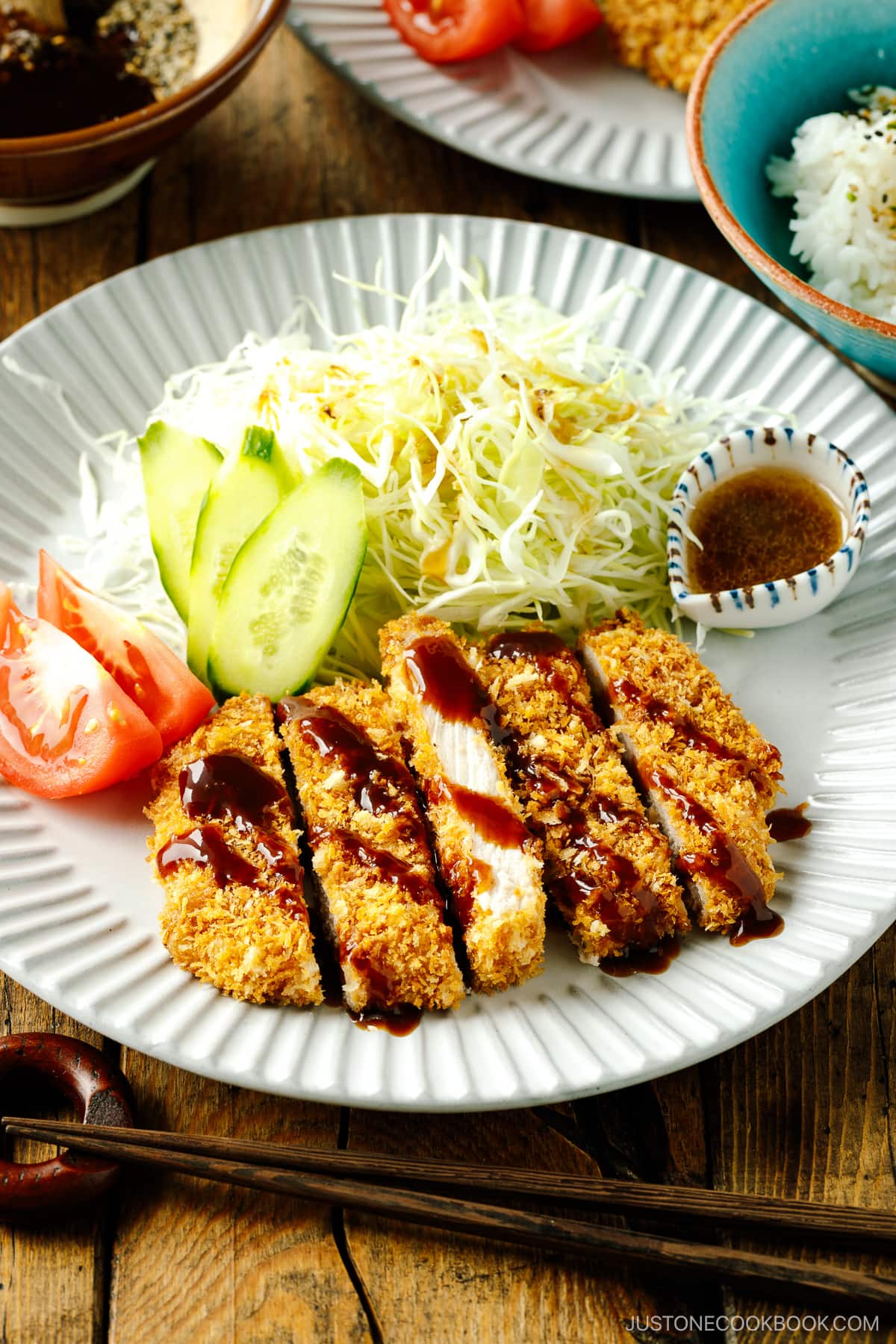 A fluted plate containing baked Tonkatsu drizzled with tonkatsu sauce, shredded cabbage, cucumber slices, and tomato wedges.