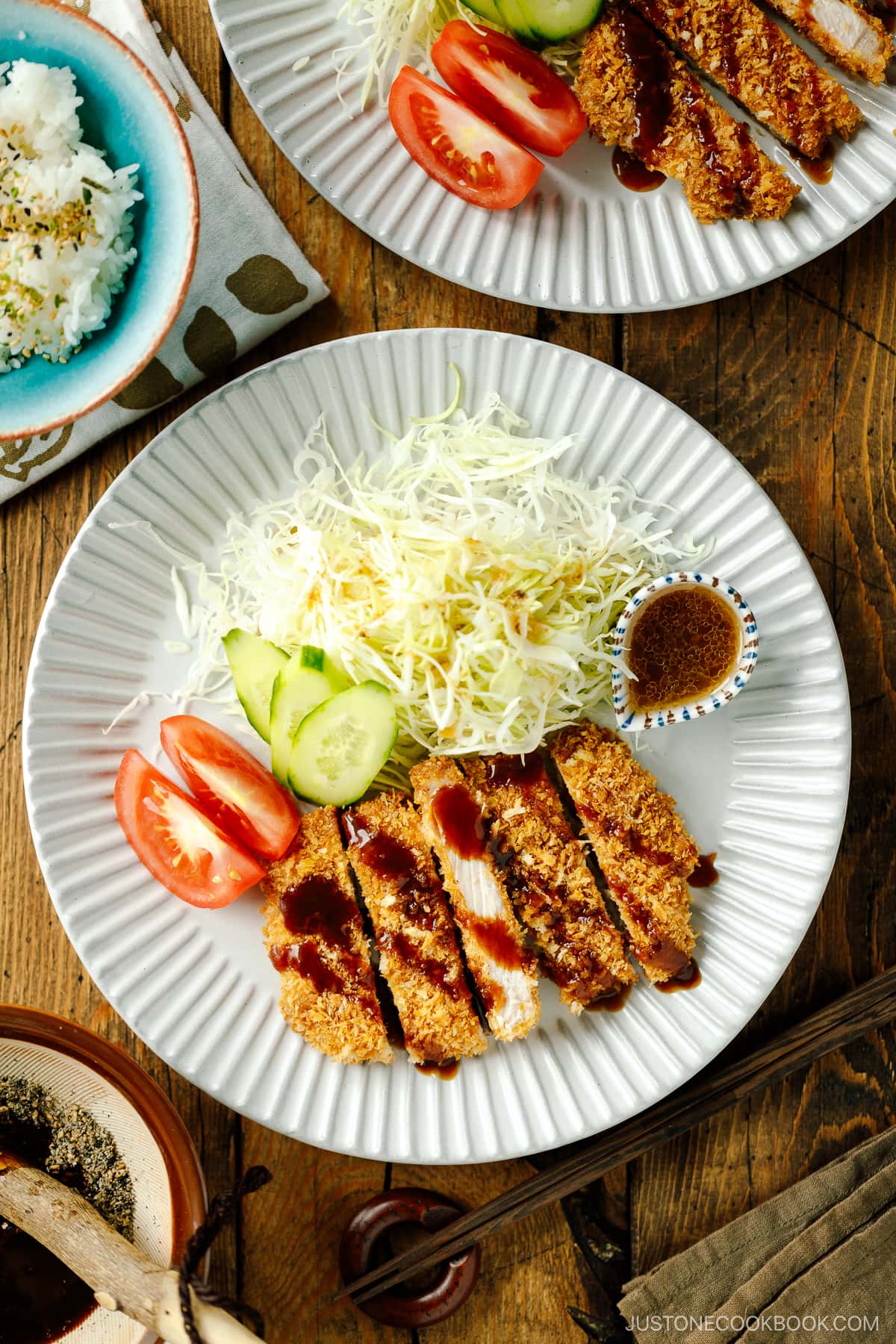 A fluted plate containing baked Tonkatsu drizzled with tonkatsu sauce, shredded cabbage, cucumber slices, and tomato wedges.