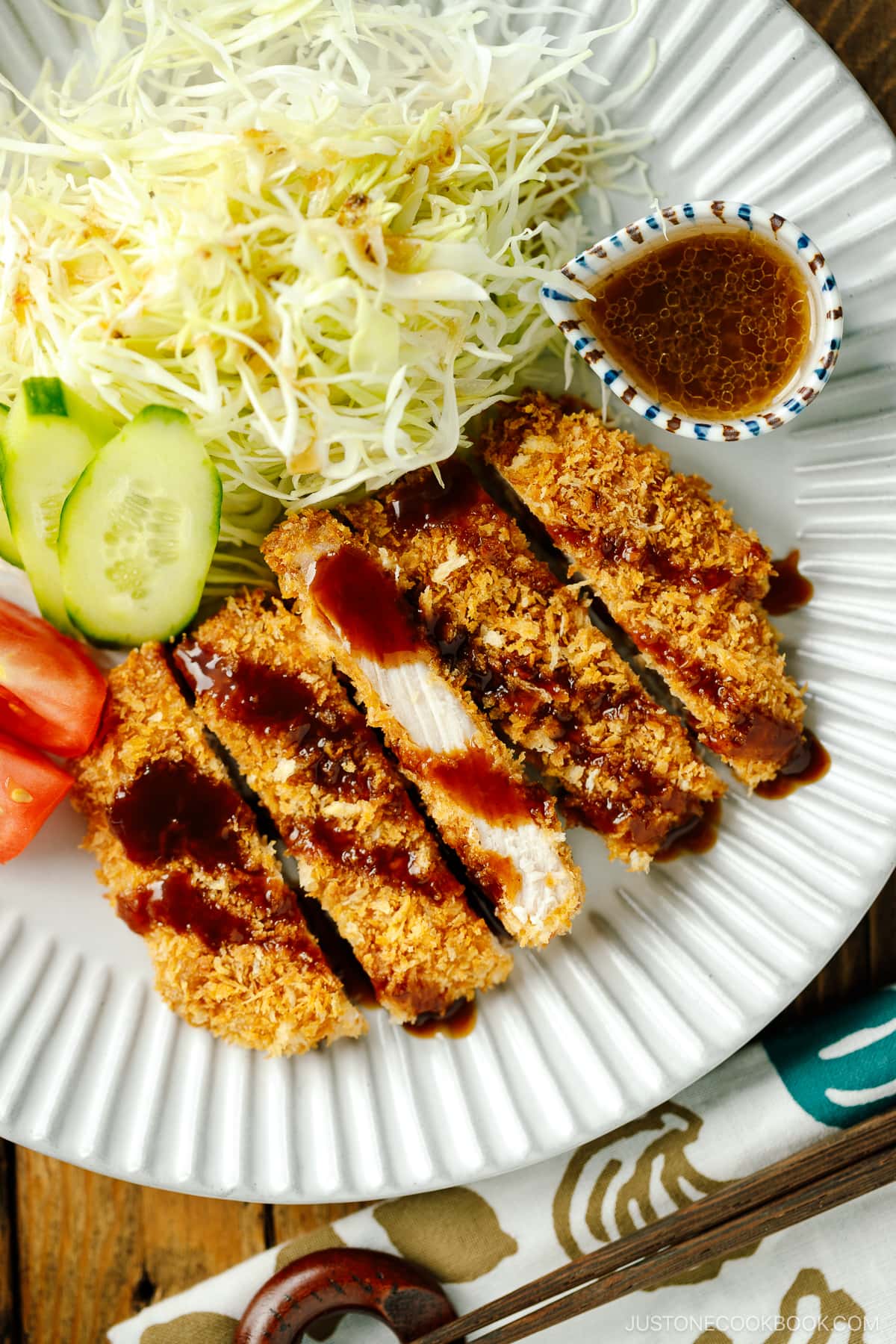 A fluted plate containing baked Tonkatsu drizzled with tonkatsu sauce, shredded cabbage, cucumber slices, and tomato wedges.