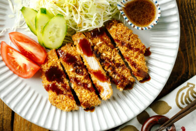 A fluted plate containing baked Tonkatsu drizzled with tonkatsu sauce, shredded cabbage, cucumber slices, and tomato wedges.