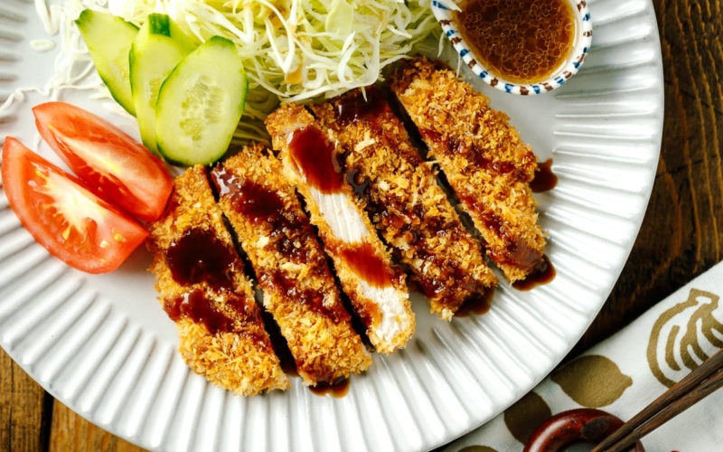 A fluted plate containing baked Tonkatsu drizzled with tonkatsu sauce, shredded cabbage, cucumber slices, and tomato wedges.