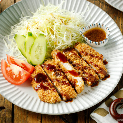 A fluted plate containing baked Tonkatsu drizzled with tonkatsu sauce, shredded cabbage, cucumber slices, and tomato wedges.