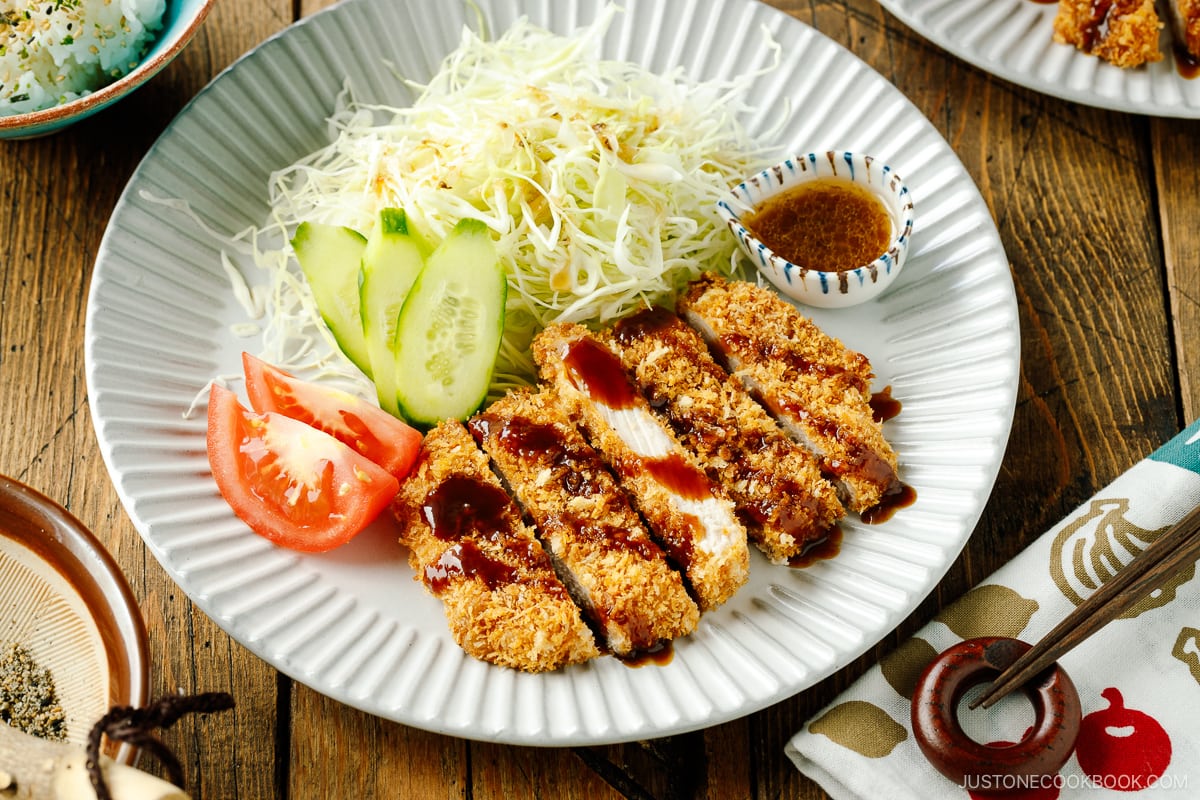 A fluted plate containing baked Tonkatsu drizzled with tonkatsu sauce, shredded cabbage, cucumber slices, and tomato wedges.