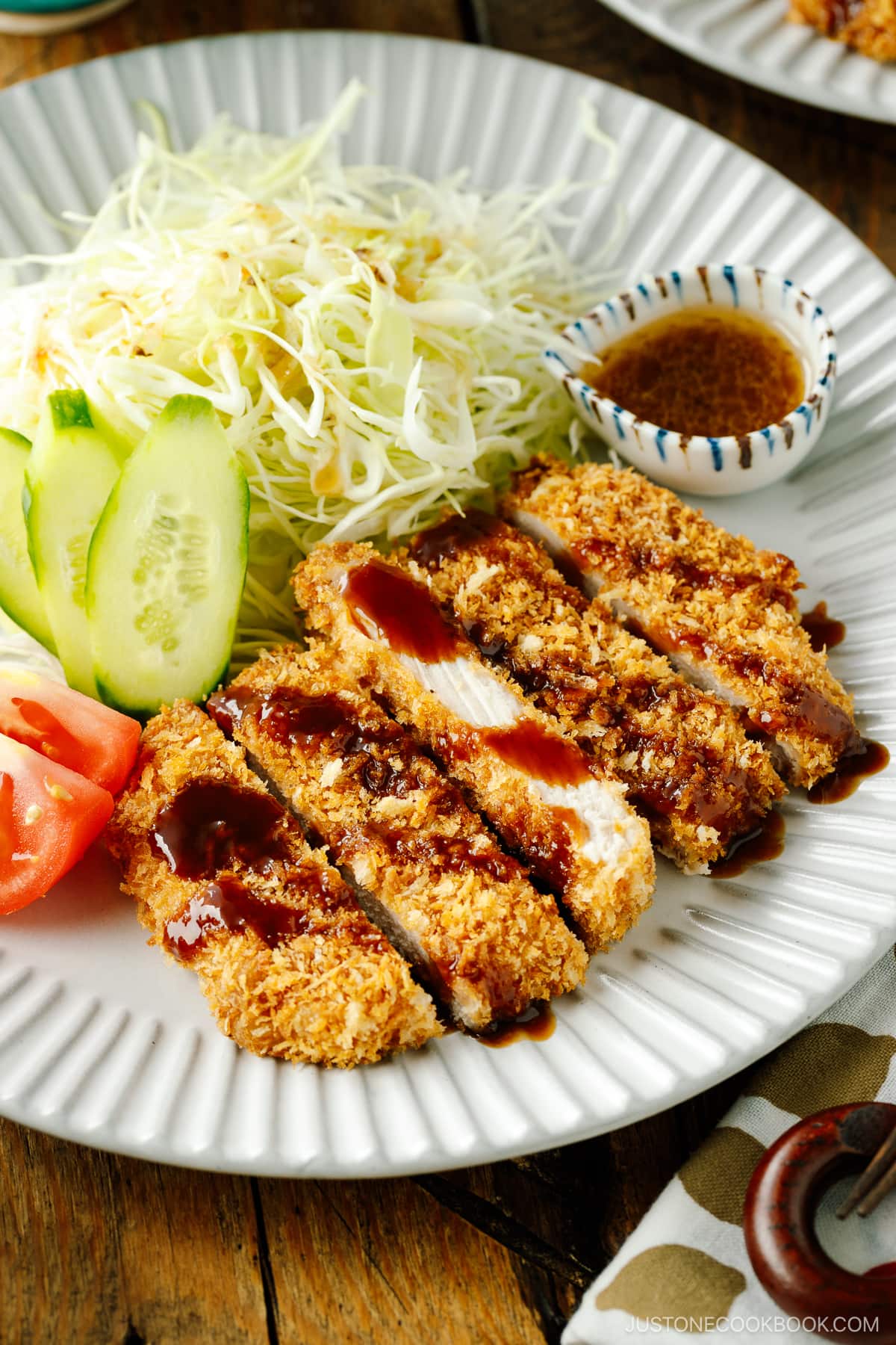 A fluted plate containing baked Tonkatsu drizzled with tonkatsu sauce, shredded cabbage, cucumber slices, and tomato wedges.