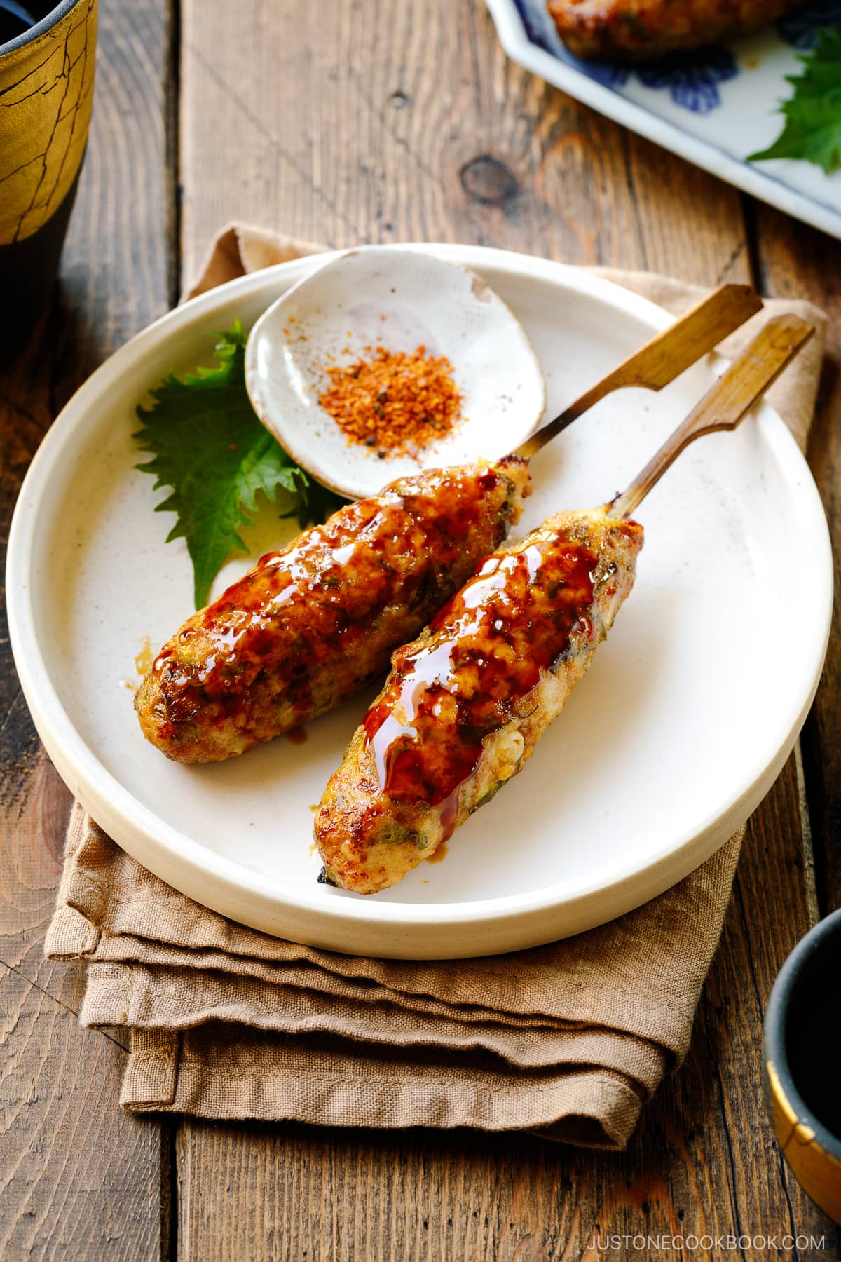 A white plate containing two skewers of Tsukune (Japanese chicken meatball) accompanied by shichimi togarashi, Japanese seven spices.
