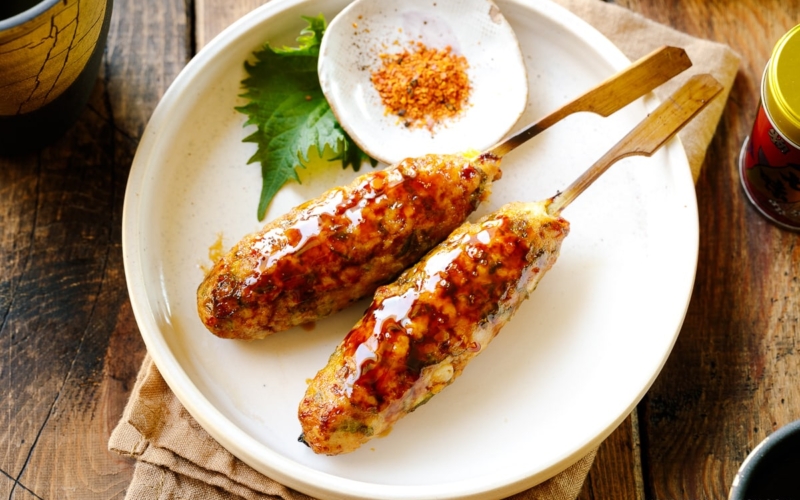 A white plate containing two skewers of Tsukune (Japanese chicken meatball) accompanied by shichimi togarashi, Japanese seven spices.