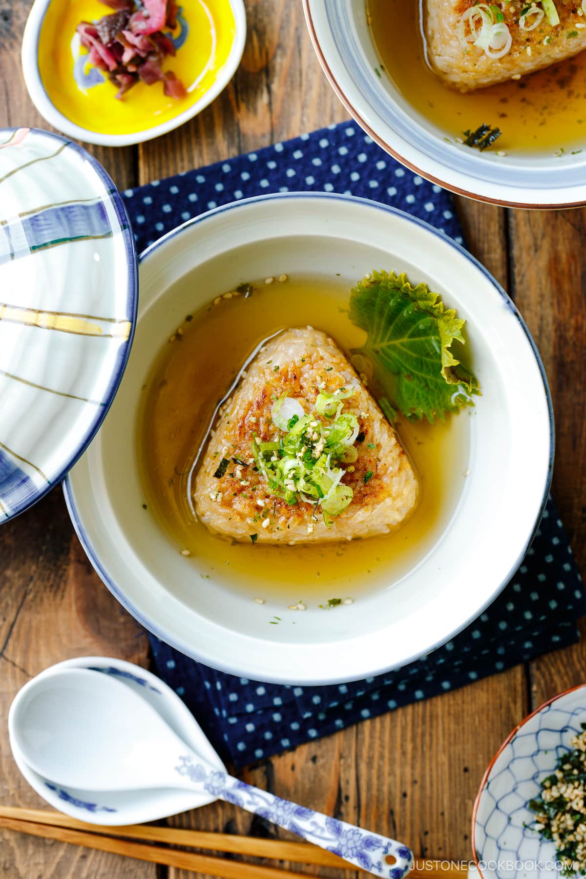 A donburi bowl containing a Japanese grilled rice ball in a flavorful dashi broth, topped with green onions and furikake rice seasoning.