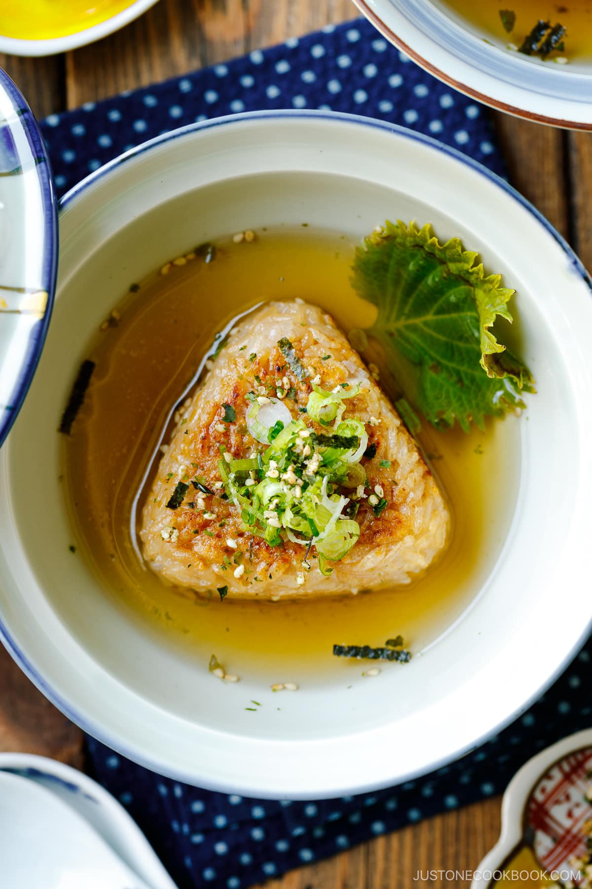 A donburi bowl containing a Japanese grilled rice ball in a flavorful dashi broth, topped with green onions and furikake rice seasoning.