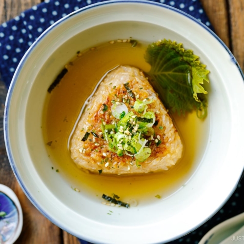 A donburi bowl containing a Japanese grilled rice ball in a flavorful dashi broth, topped with green onions and furikake rice seasoning.