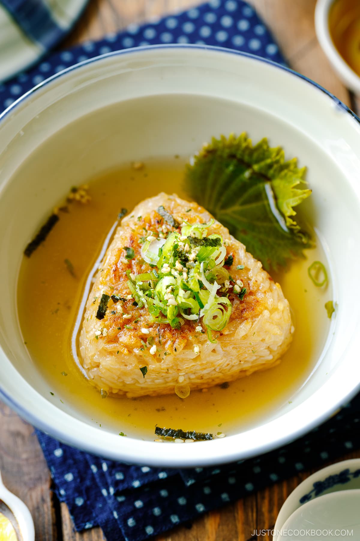A donburi bowl containing a Japanese grilled rice ball in a flavorful dashi broth, topped with green onions and furikake rice seasoning.
