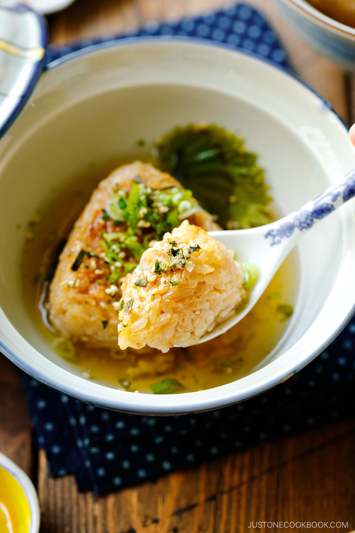 A donburi bowl containing a Japanese grilled rice ball in a flavorful dashi broth, topped with green onions and furikake rice seasoning.