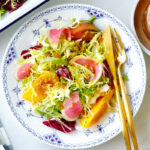 A white and blue plate containing frisee and radicchio lettuce, thinly sliced watermelon radish, and heirloom tomatoes, drizzled with Japanese onion dressing.