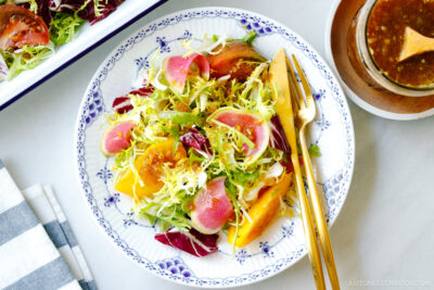 A white and blue plate containing frisee and radicchio lettuce, thinly sliced watermelon radish, and heirloom tomatoes, drizzled with Japanese onion dressing.