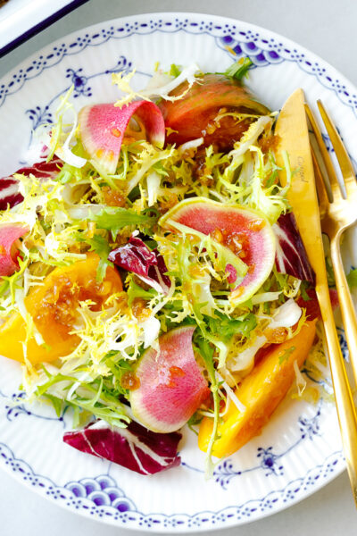 A white and blue plate containing frisee and radicchio lettuce, thinly sliced watermelon radish, and heirloom tomatoes, drizzled with Japanese onion dressing.