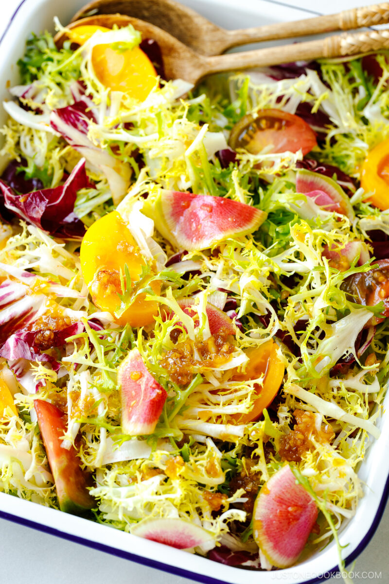 A white and blue enamel tray containing frisee and radicchio lettuce, thinly sliced watermelon radish, and heirloom tomatoes, drizzled with Japanese onion dressing.