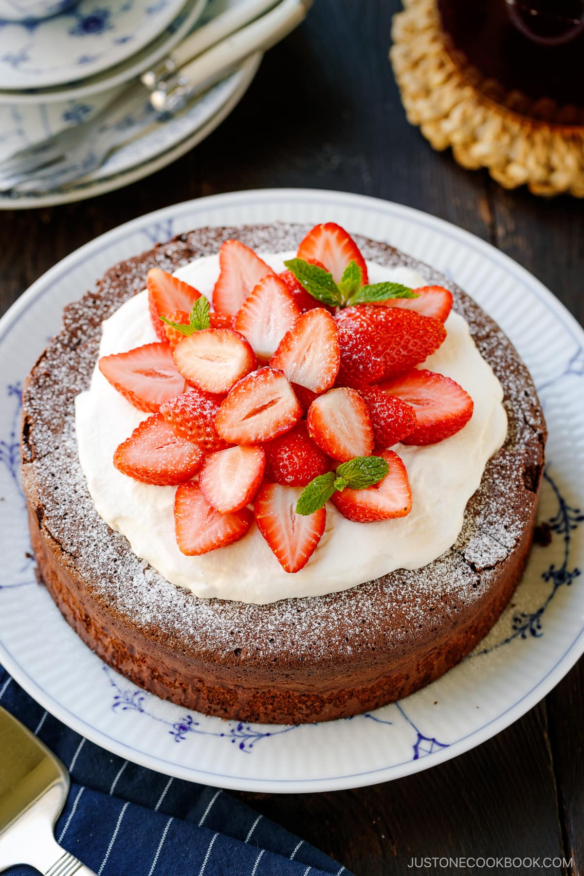A large plate containing Chocolate Gateau (Chocolate Cake) dusted with powdered sugar and decorated with whipped cream and strawberries.