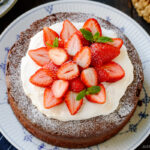 A large plate containing Chocolate Gateau (Chocolate Cake) dusted with powdered sugar and decorated with whipped cream and strawberries.