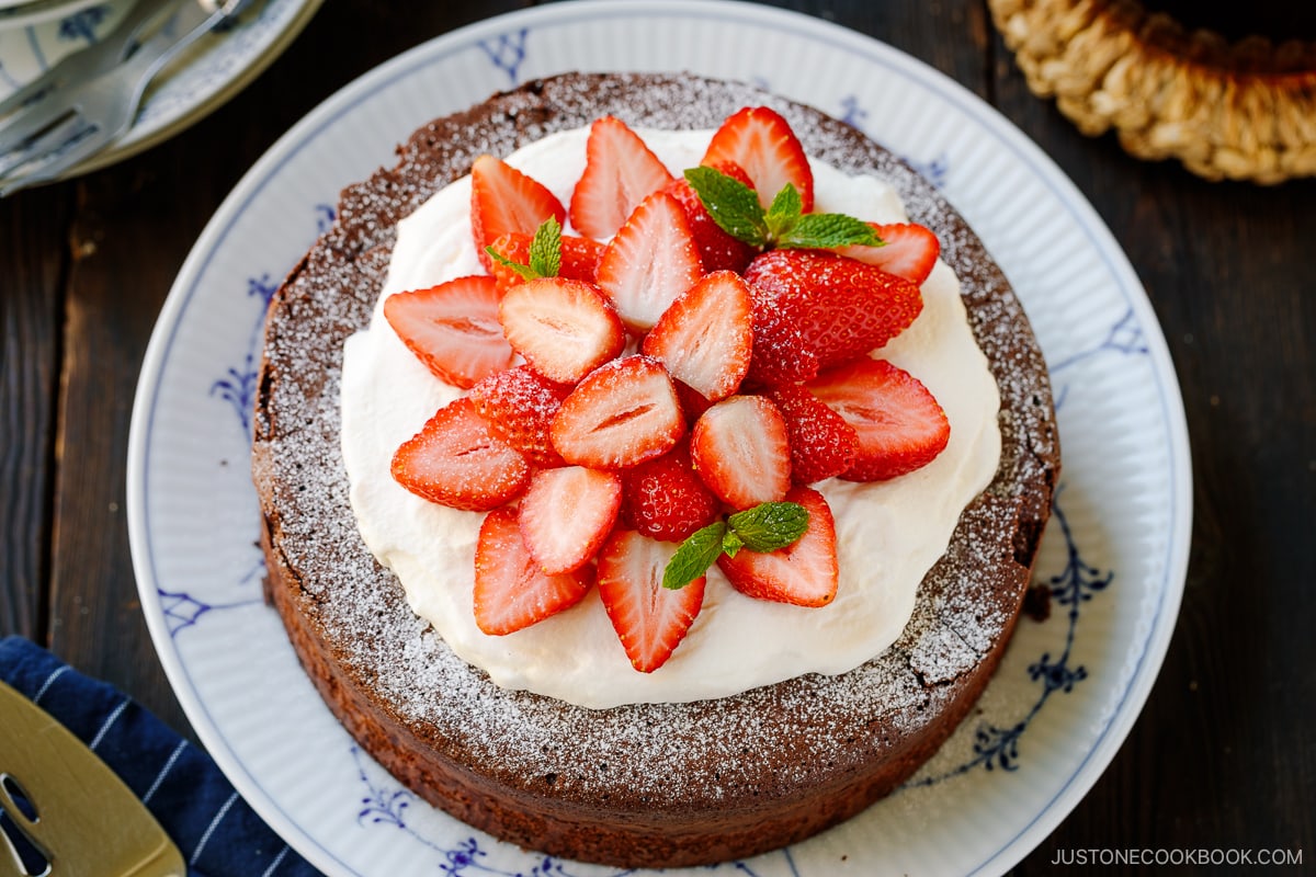 A large plate containing Chocolate Gateau (Chocolate Cake) dusted with powdered sugar and decorated with whipped cream and strawberries.