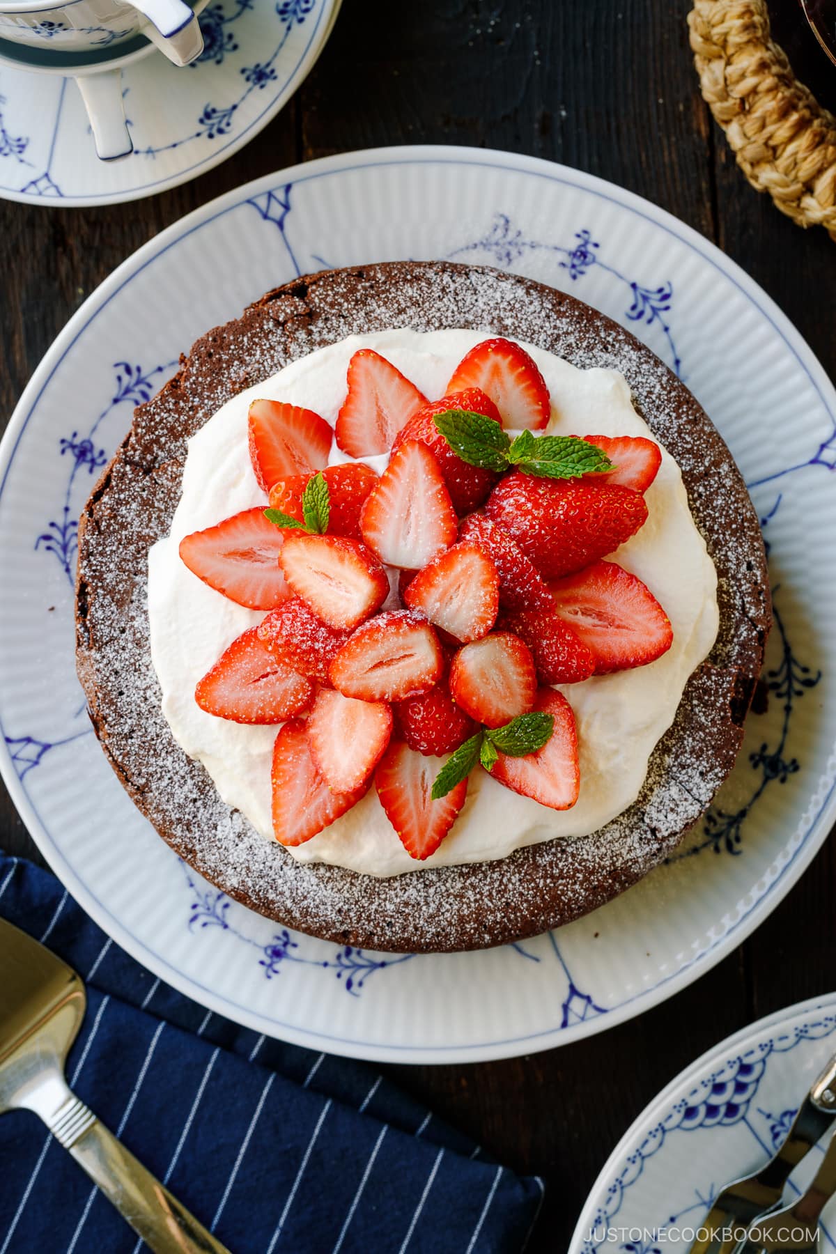 A large plate containing Chocolate Gateau (Chocolate Cake) dusted with powdered sugar and decorated with whipped cream and strawberries.