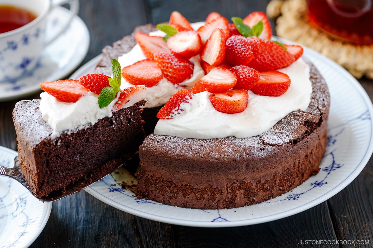 A large plate containing Chocolate Gateau (Chocolate Cake) dusted with powdered sugar and decorated with whipped cream and strawberries.