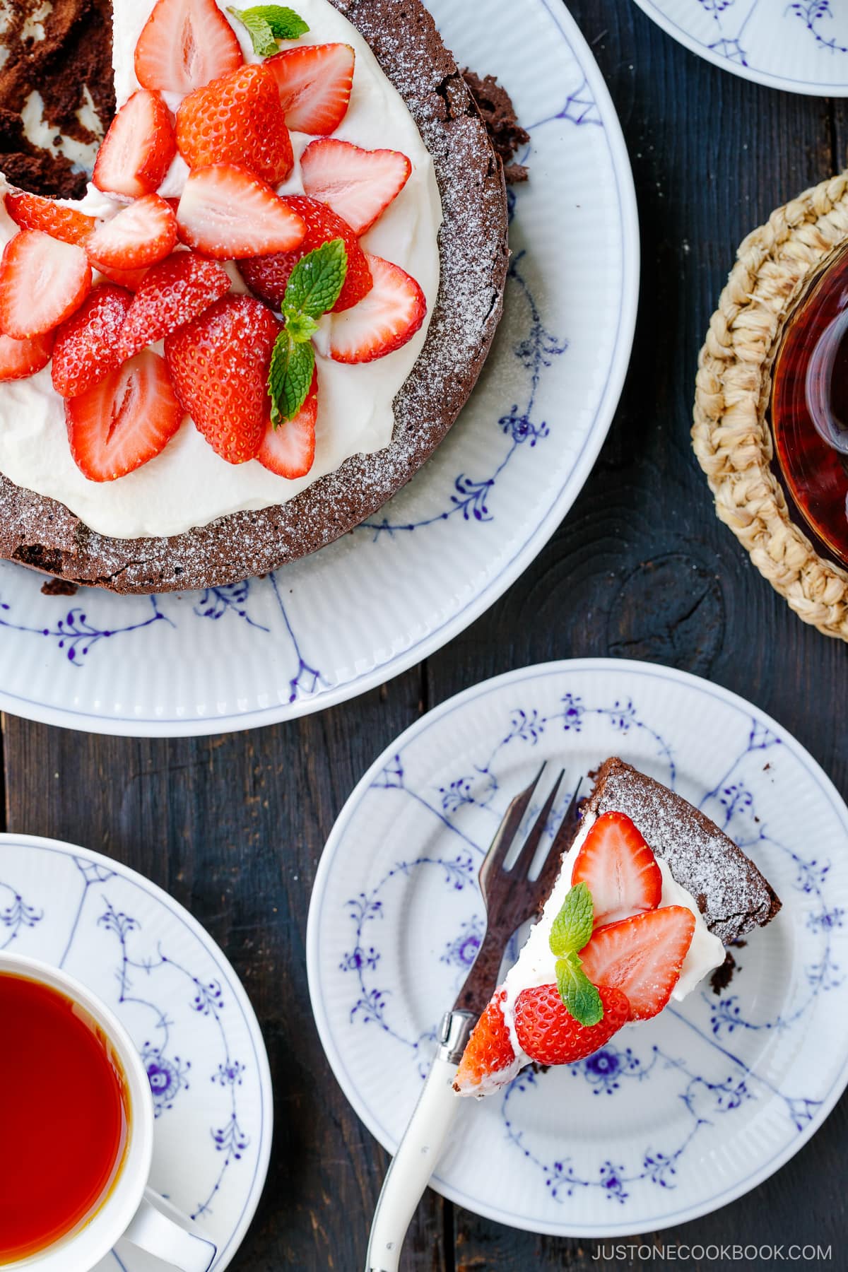 A plate containing a slice of Chocolate Gateau (Chocolate Cake) topped with whipped cream and strawberry slices and the remaining cake on the side.