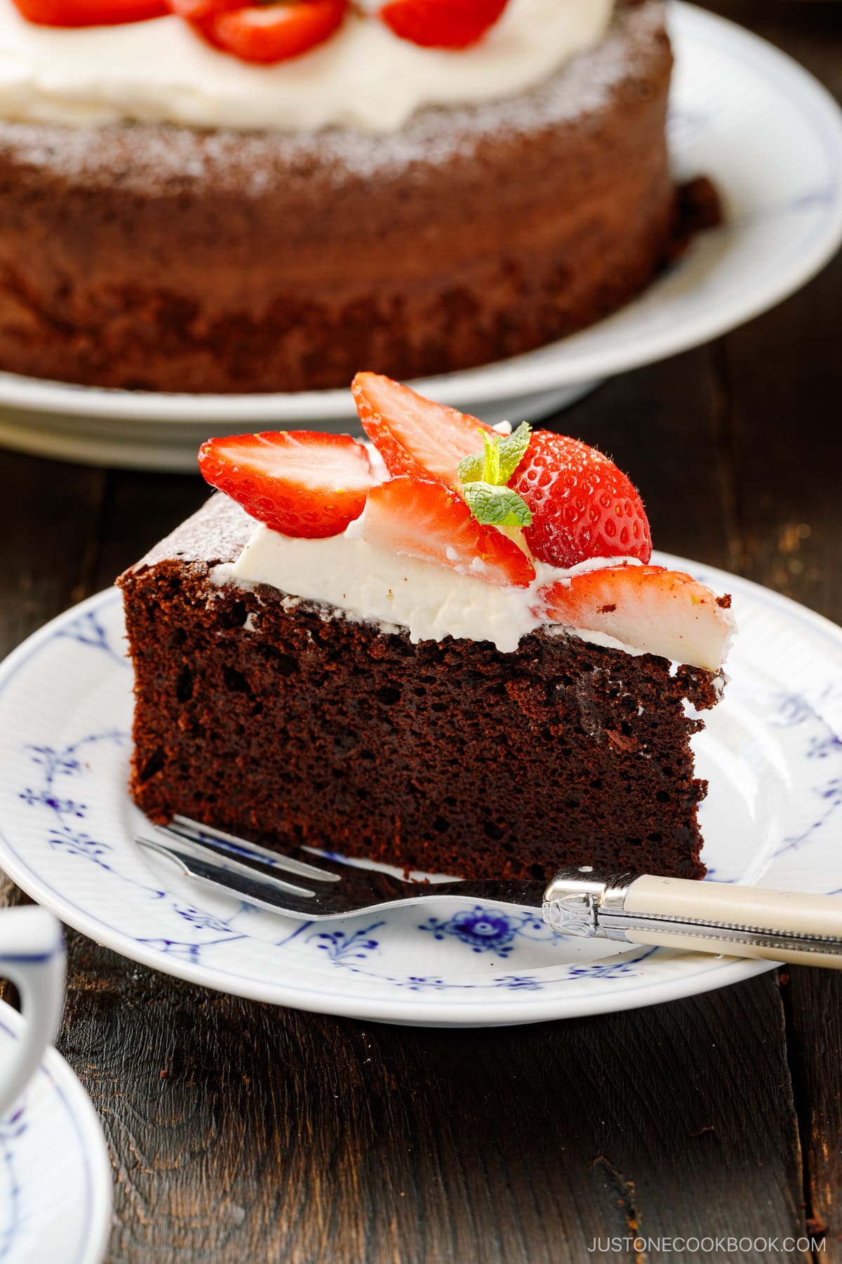 A plate containing a slice of Chocolate Gateau (Chocolate Cake), topped with whipped cream and strawberry slices.