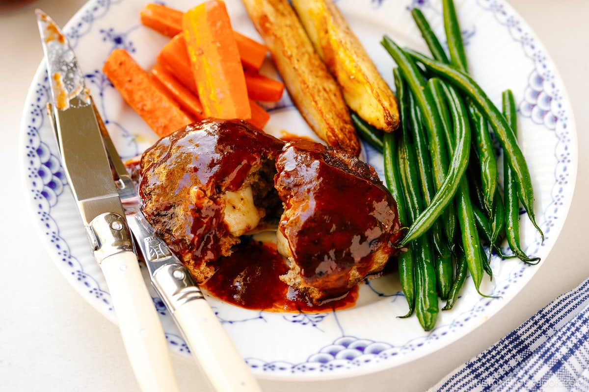 A white and blue plate containing Cheese Stuffed Japanese Hamburger Steak (Hambagu) stuffed with cheese and slathered in demi-glace sauce.