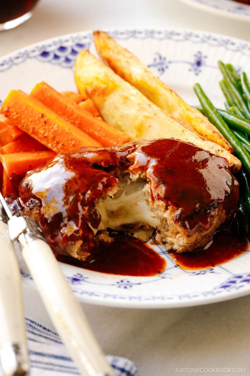 A white and blue plate containing Cheese Stuffed Japanese Hamburger Steak (Hambagu) stuffed with cheese and slathered in demi-glace sauce.