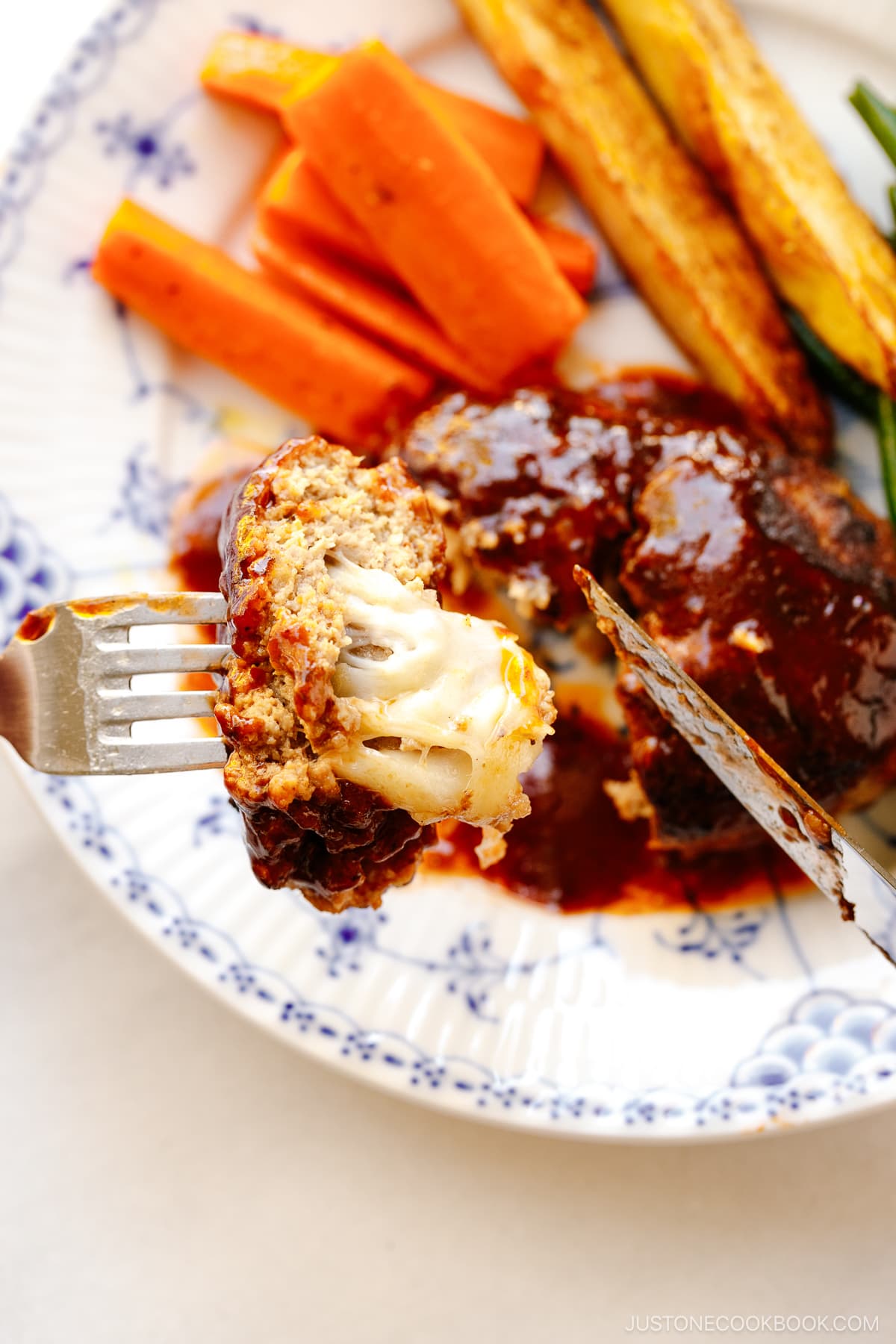 A white and blue plate containing Cheese Stuffed Japanese Hamburger Steak (Hambagu) stuffed with cheese and slathered in demi-glace sauce.