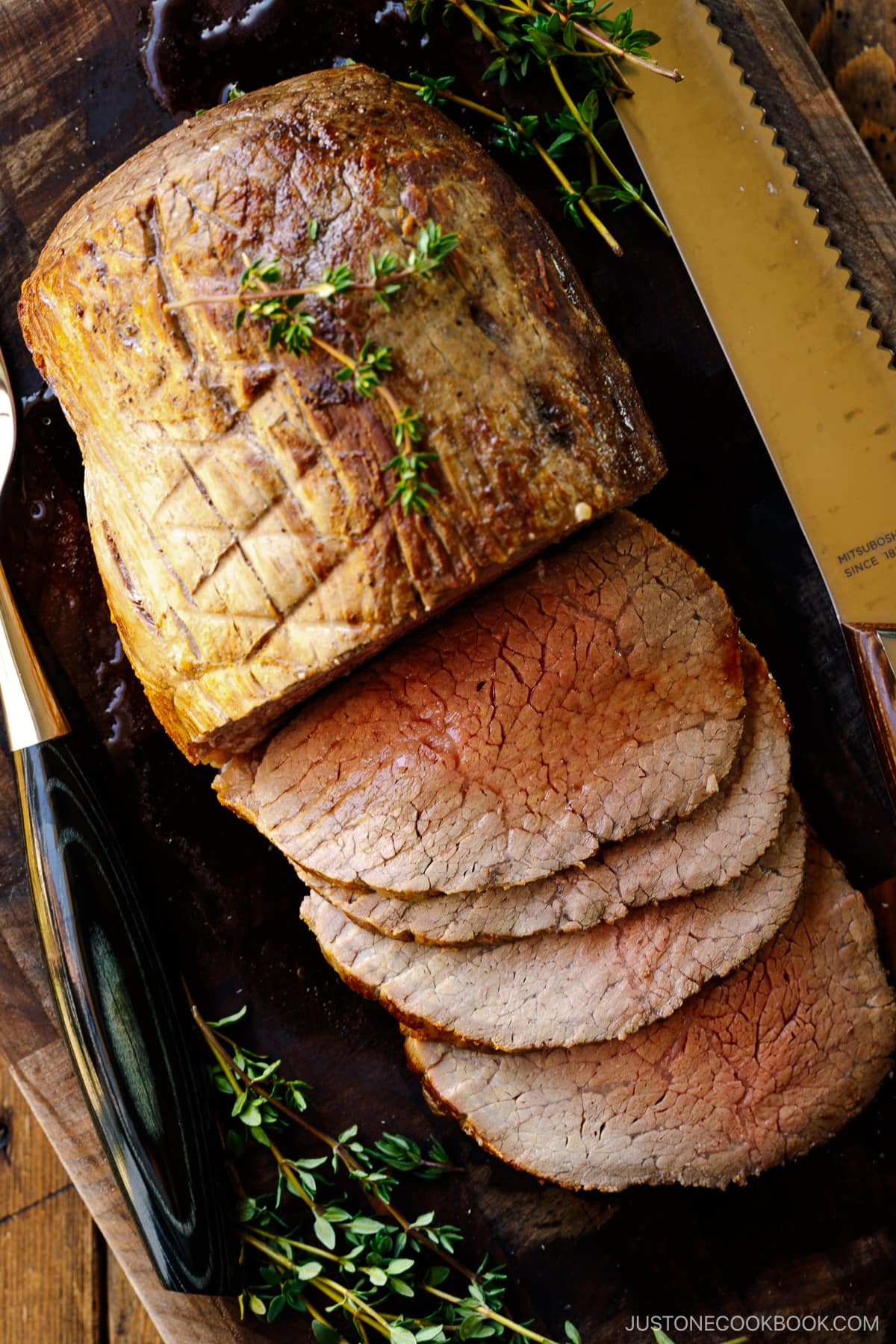 A roast beef being thinly sliced on the wooden cutting board garnished with thyme.