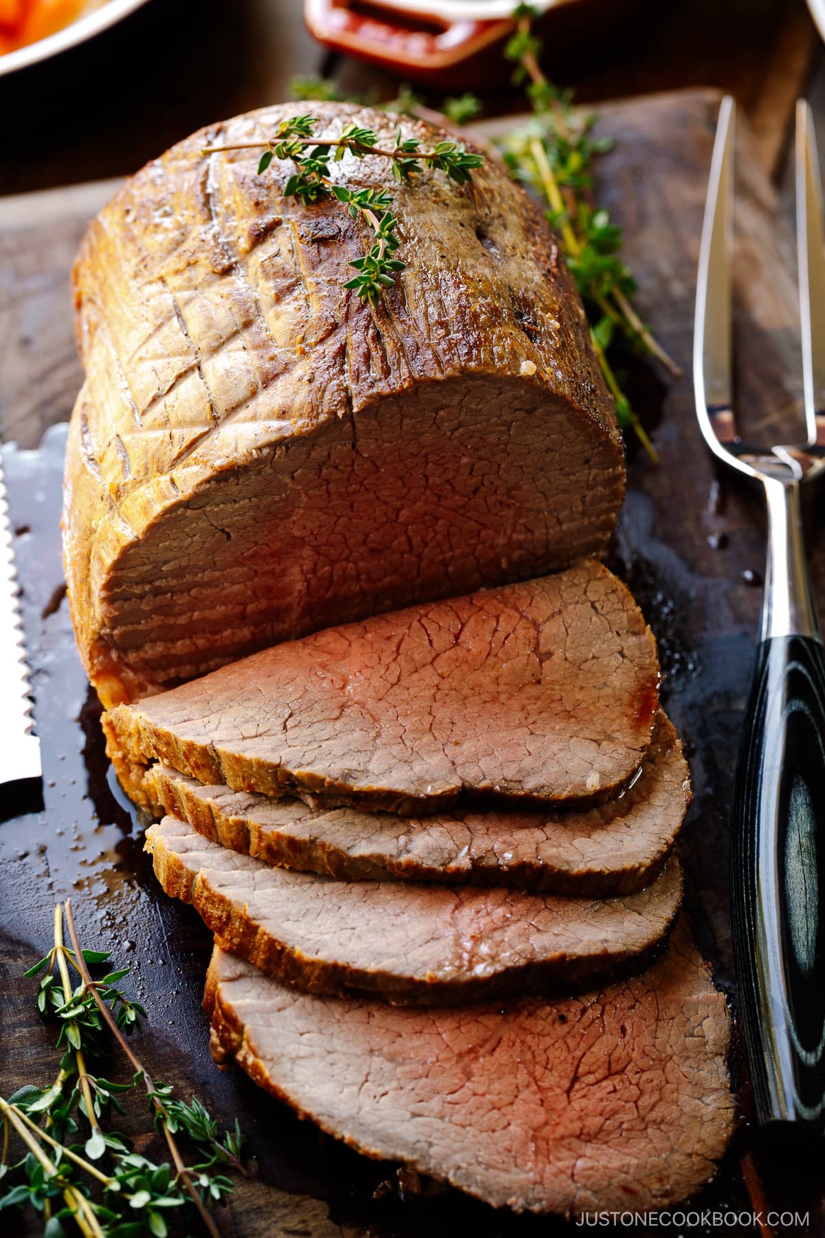 A roast beef being thinly sliced on the wooden cutting board garnished with thyme.