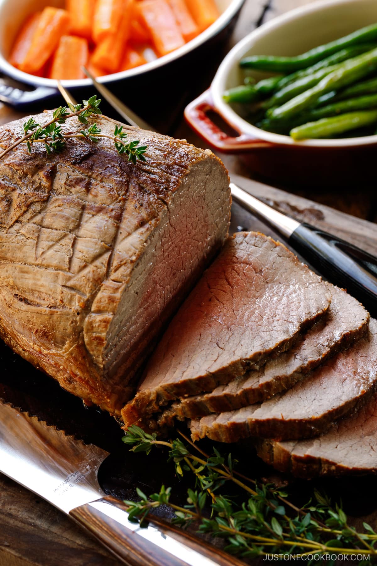 A roast beef being thinly sliced on the wooden cutting board garnished with thyme.