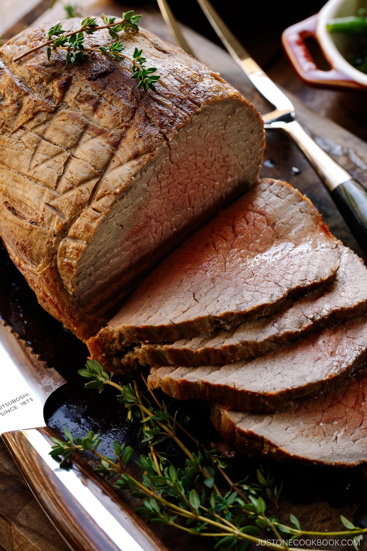 A roast beef being thinly sliced on the wooden cutting board garnished with thyme.