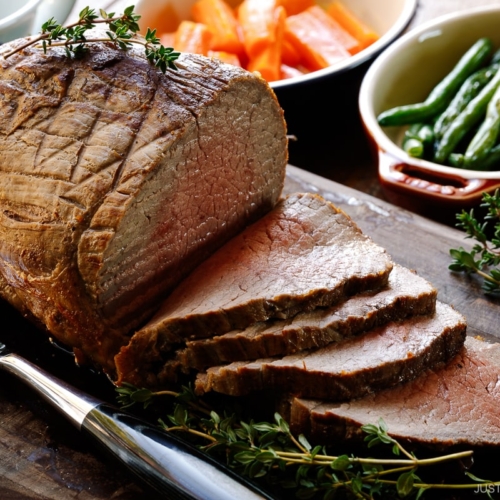 A roast beef being thinly sliced on the wooden cutting board garnished with thyme.