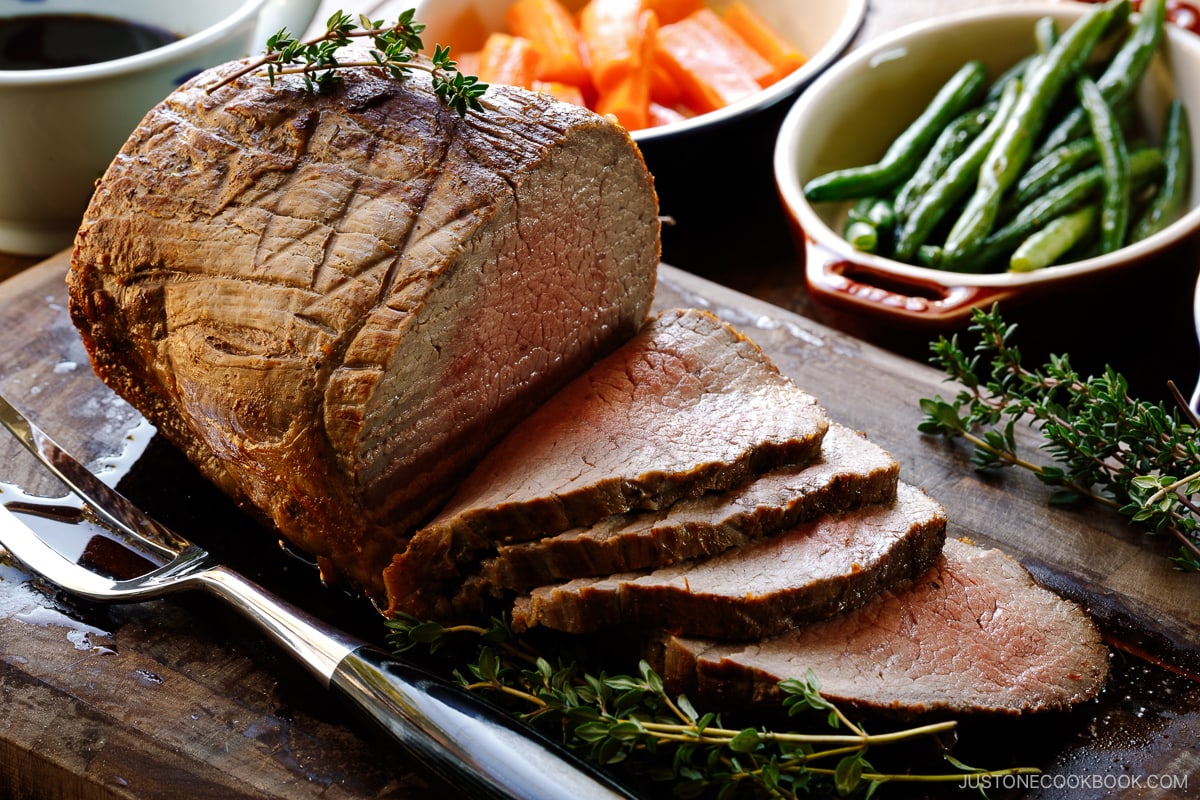 A roast beef being thinly sliced on the wooden cutting board garnished with thyme.